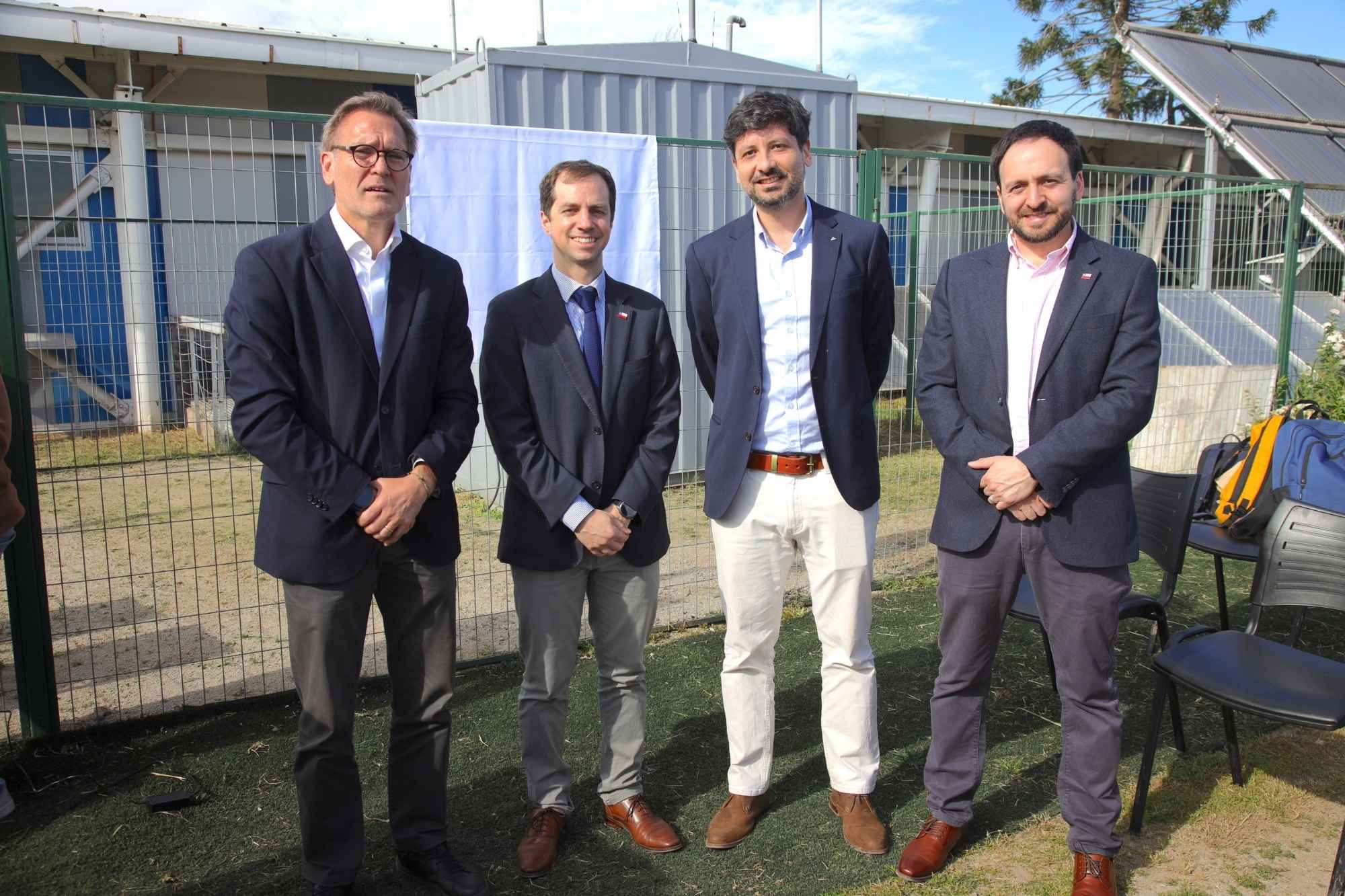 Cuatro hombres de traje sonrientes junto a vallas y paneles solares, fotografía al aire libre